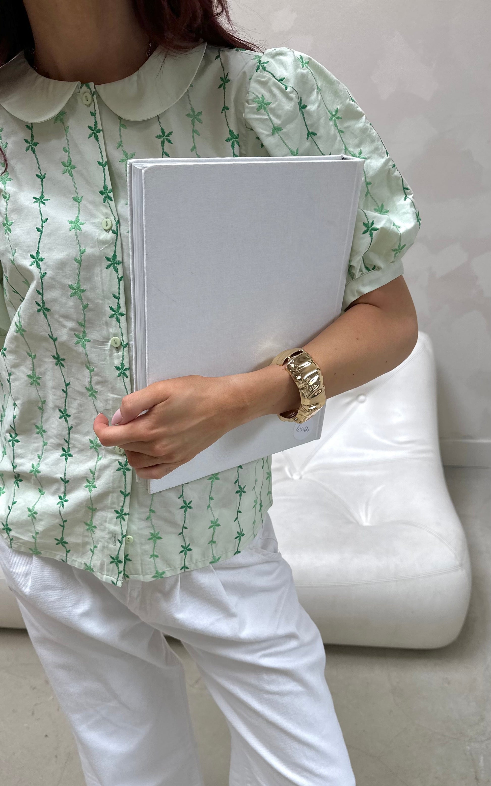 Person wearing a green patterned shirt and white pants holding a white folder.