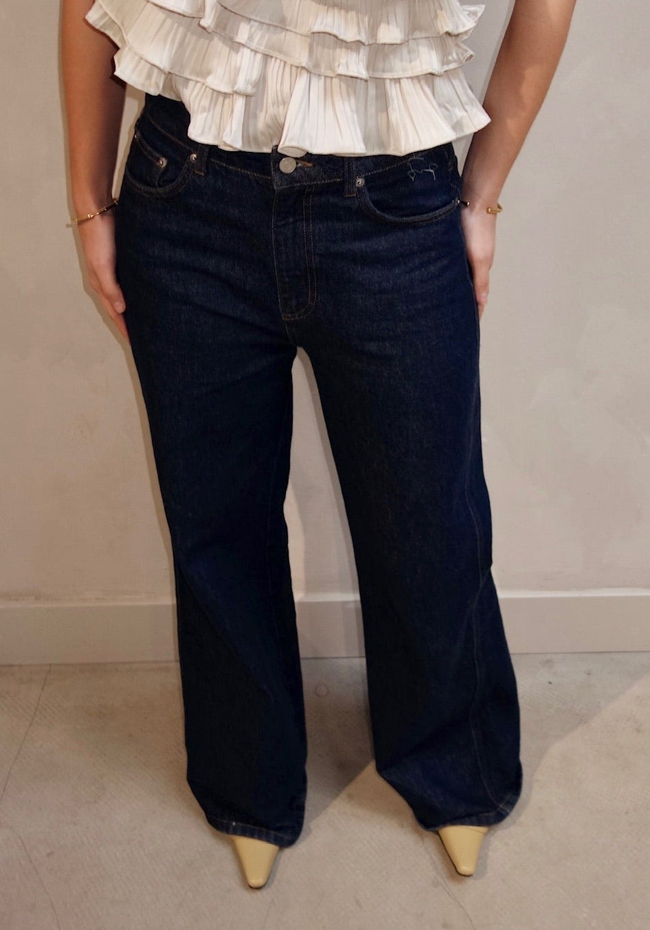 Woman wearing a ruffled blouse and dark jeans against a plain background