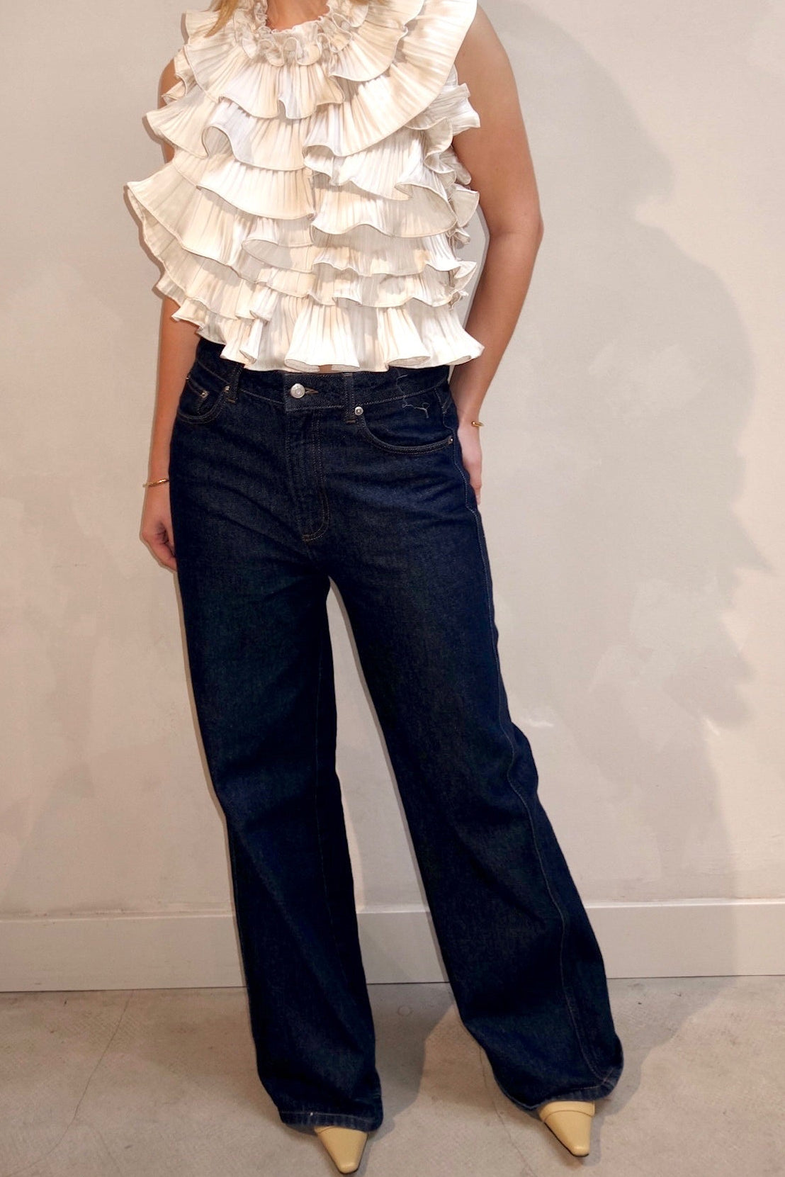 Woman wearing a white ruffled top and dark blue jeans against a plain background