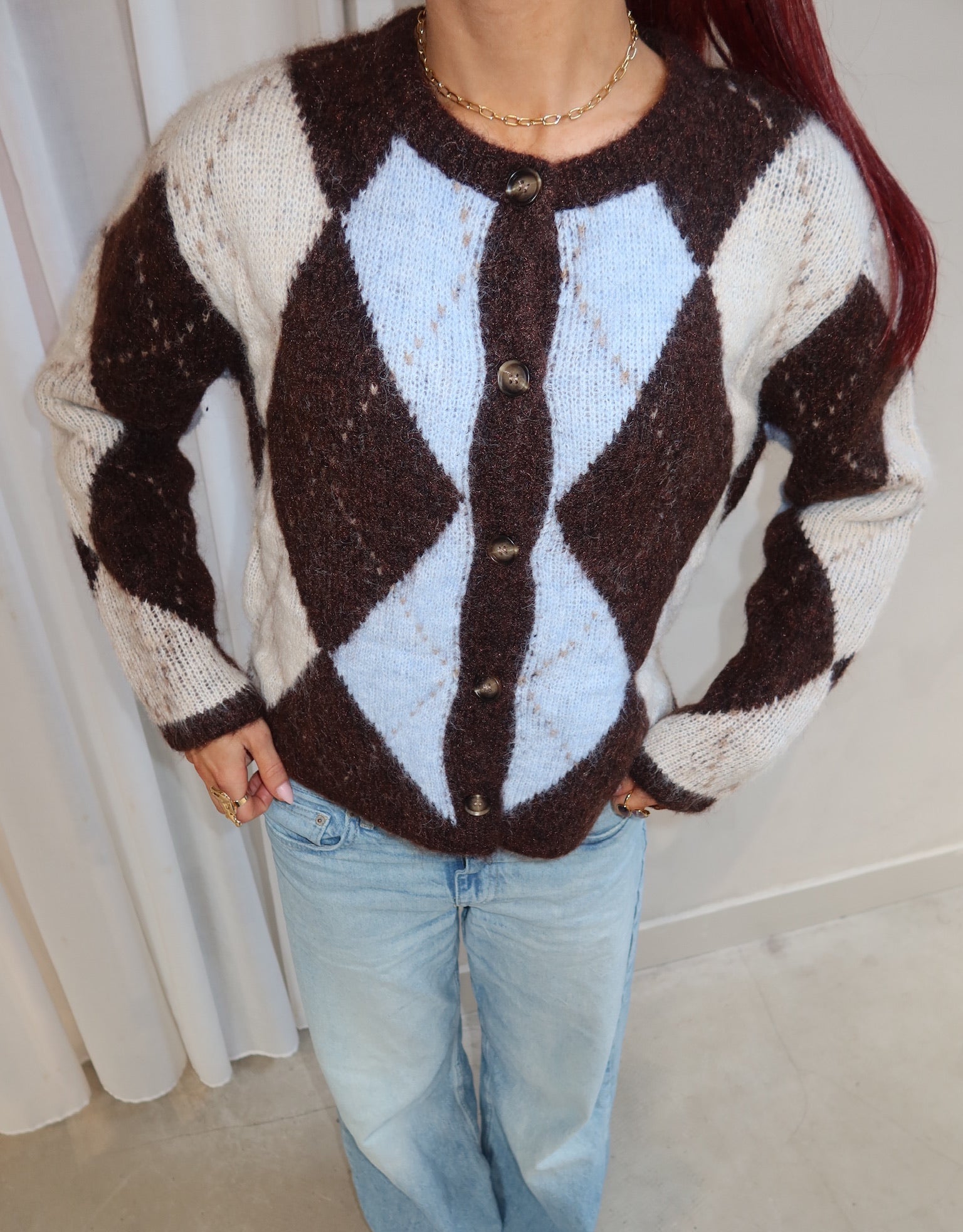 Person wearing a brown and white argyle patterned cardigan with light blue jeans indoors.