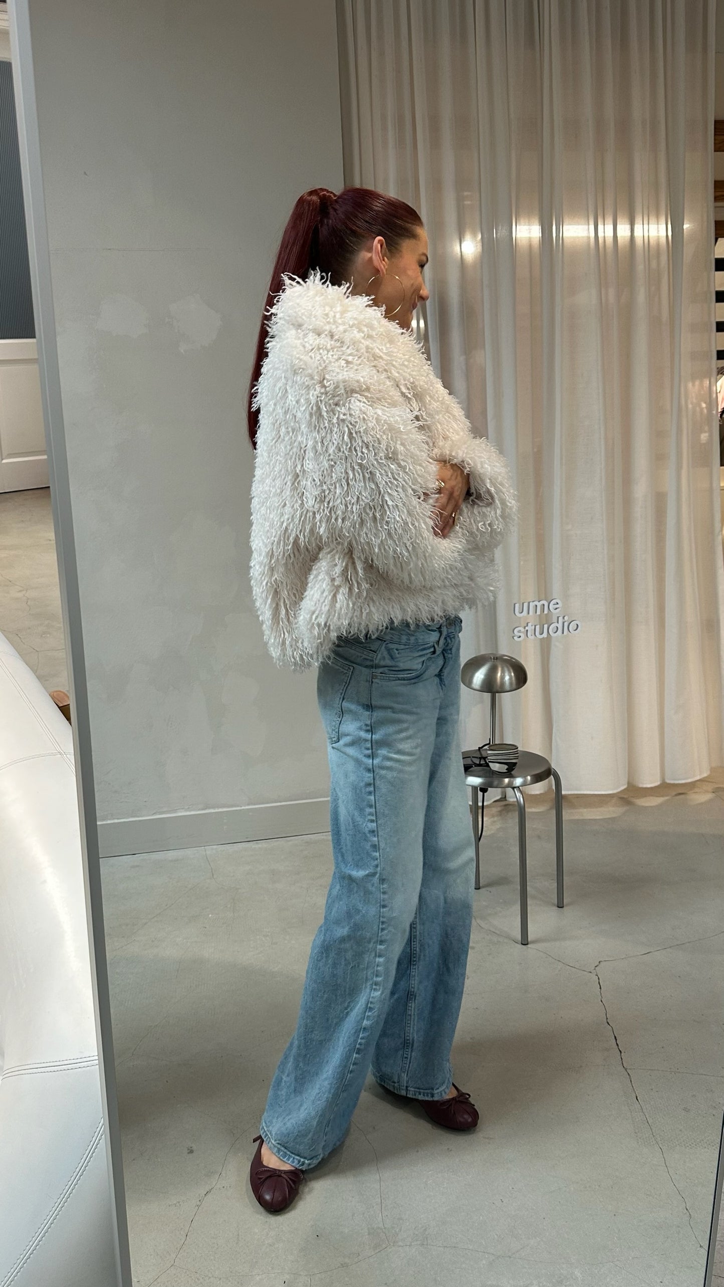 Person wearing a fluffy white jacket and blue jeans standing in front of a mirror.